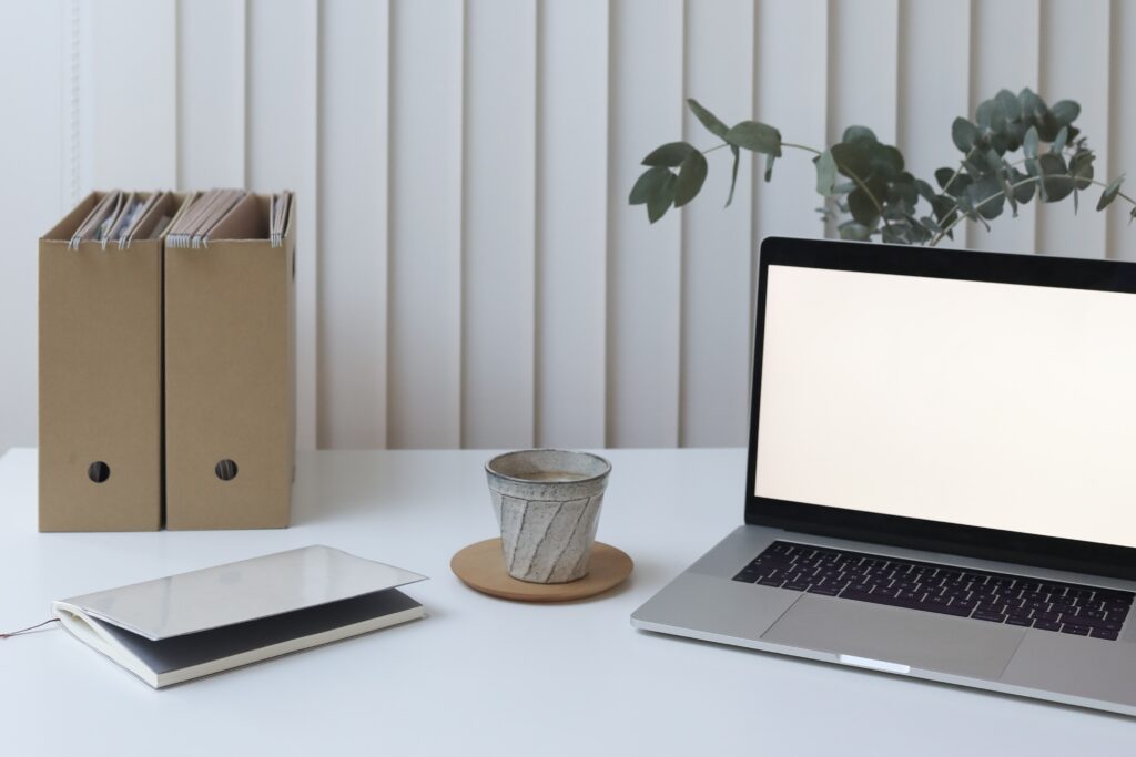 an open laptop on a white desk with a plant behind it, a cup of coffee on saucer to the side and notebook